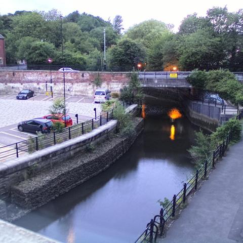 Photograph of the tidal stretch of Ouseburn River at south side of Ouseburn Bridge in Newcastle upon Tyne. To the left (south-west) of the bridge is Cut Bank, and to the right (north-east) is Byker Bank. In the foreground is the pedestrian Riverside Walk and the photo also spans some surrounding built-up areas and trees, with sky in the background. Still image captured from webcam at 21:44 hrs on Friday 4 July 2025.