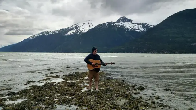 John Swanke plays guitar on a rocky shore with snow-capped mountains and cloudy sky in the background, creating a serene and moody scene.
