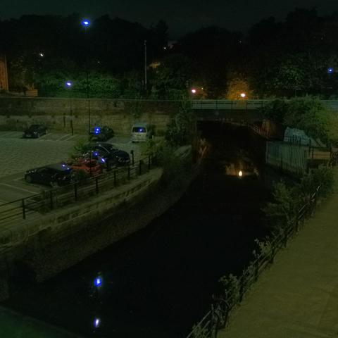 Photograph of the tidal stretch of Ouseburn River at south side of Ouseburn Bridge in Newcastle upon Tyne. To the left (south-west) of the bridge is Cut Bank, and to the right (north-east) is Byker Bank. In the foreground is the pedestrian Riverside Walk and the photo also spans some surrounding built-up areas and trees, with sky in the background. Still image captured from webcam at 23:44 hrs on Friday 4 July 2025.