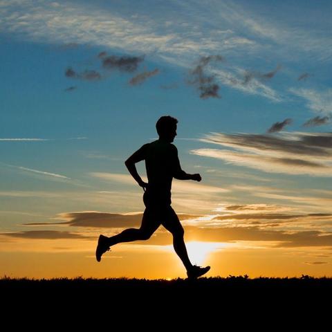 Photograph of a man running, seen in silhouette, against a yellow sunset. The blue of the sky above the horizon is dotted with tiny clouds.