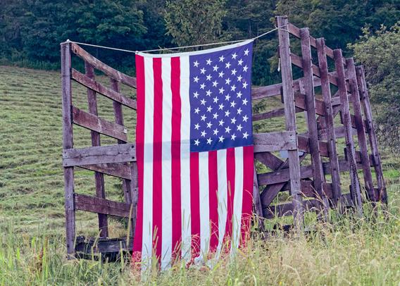 An American flag hangs from a line strung across the back of an old wooden hay wagon with grass in front and rows of cut grass in a field in the back