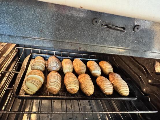 12 potatoes on a sheet pan in an oven. The potatoes are thinly sliced without being sliced all the way through, hasselback style. They are halfway done cooking, so they are starting to fan out along the slices