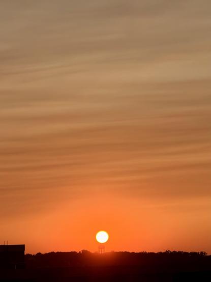 The sky is lit up by a setting sun with light clouds and an orange red gradient. The sun looks like a perfect circle just above the horizon. It is almost exactly hovering over a water tower. 