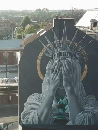 Photo of a new mural by Dutch artist Judith de Leeuw on the side of a building in Roubaix, France. It shows the Statue of Liberty holding her hands up against her face as if she is crying into them. There are other buildings behind and beside the building with the mural, as well as many green treetops.