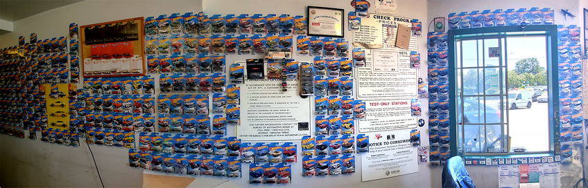 Panorama of the interior of a small smog testing station with the walls completely covered with Hot Wheels toy cars, still sealed in their original packages.