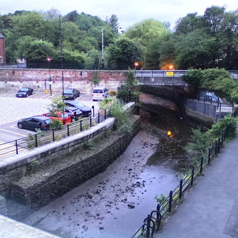 Photograph of the tidal stretch of Ouseburn River at south side of Ouseburn Bridge in Newcastle upon Tyne. To the left (south-west) of the bridge is Cut Bank, and to the right (north-east) is Byker Bank. In the foreground is the pedestrian Riverside Walk and the photo also spans some surrounding built-up areas and trees, with sky in the background. Still image captured from webcam at 04:32 hrs on Saturday 5 July 2025.