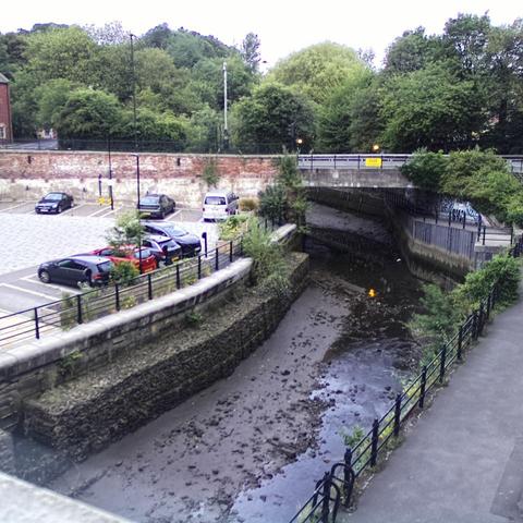 Photograph of the tidal stretch of Ouseburn River at south side of Ouseburn Bridge in Newcastle upon Tyne. To the left (south-west) of the bridge is Cut Bank, and to the right (north-east) is Byker Bank. In the foreground is the pedestrian Riverside Walk and the photo also spans some surrounding built-up areas and trees, with sky in the background. Still image captured from webcam at 06:00 hrs on Saturday 5 July 2025.
