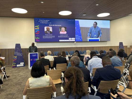 A conference scene showing a speaker addressing an audience at the Francophone Forum on Internet Governance and AI. The stage features two speakers' portraits and presentation materials. Various attendees are seated, engaged in the event.