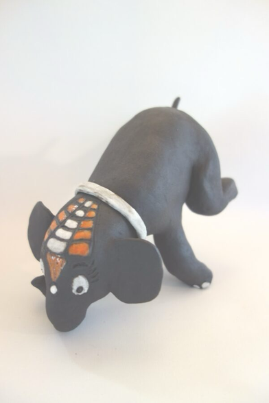 Elefant aus schwarzem Ton, der auf dem Rüssel steht und beide Hinterbeine in die Luft streckt. Er hat ein weißes Halsband und die Stirn ist mit einem weiß—orangen Muster bemalt.