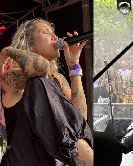 Das Bild zeigt eine Frau (Kim), die auf einer Bühne singt. Sie hat blonde, wellige Haare und ist mit zahlreichen Tattoos bedeckt, darunter ein Adler auf ihrem Arm und ein Blitzmuster. Sie trägt ein schwarzes Oberteil und eine schwarze Hose mit einem elastischen Bund. Ihre Hand, die einen Mikrofonständer hält, ist mit roten Nagellack bemalt. Sie hat ein Armband mit einem violetten Band am Handgelenk. Im Hintergrund sieht man eine Menge von Menschen, die sie ansehen und applaudieren, einige mache…