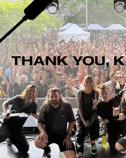 Das Bild zeigt eine Gruppe von Musikern auf einer Bühne, die eine große, jubelnde Menge hinter sich sieht. Die Musiker sind in der Vordergrundpositioniert und tragen schwarze Kleidung. Einige von ihnen machen die "Rock on"-Geste mit ihren Händen. Die Menge hinter ihnen ist dicht gedrängt und zeigt ebenfalls die "Rock on"-Geste, während sie die Musiker anfeuert. Im Hintergrund sind weiße Zelte und Bäume zu sehen, was auf eine offene Veranstaltung hindeutet. Über dem Bild steht in großen, schwarz…