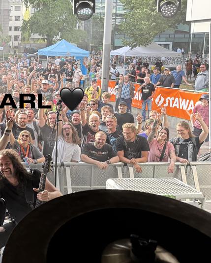 
Das Bild zeigt eine lebhafte Szene auf einer öffentlichen Bühne, wahrscheinlich während eines Musikfests. Im Vordergrund ist ein Musiker mit langen, dunklen Haaren zu sehen, der eine Gitarre spielt. Er trägt ein schwarzes T-Shirt und hat einen konzentrierten Ausdruck im Gesicht. Hinter ihm steht ein Metallzaun, durch den eine große Menschenmenge sichtbar ist. Die Zuschauer sind in einer Mischung aus Freude und Energie, viele von ihnen machen die berühmte "Rock on"-Geste mit ihren Händen. Viele…