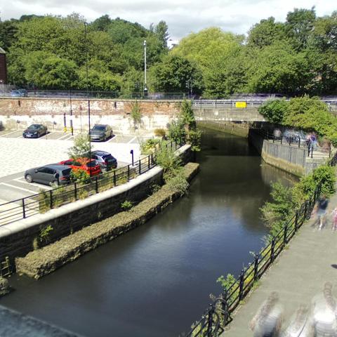 Photograph of the tidal stretch of Ouseburn River at south side of Ouseburn Bridge in Newcastle upon Tyne. To the left (south-west) of the bridge is Cut Bank, and to the right (north-east) is Byker Bank. In the foreground is the pedestrian Riverside Walk and the photo also spans some surrounding built-up areas and trees, with sky in the background. Still image captured from webcam at 12:04 hrs on Saturday 5 July 2025.