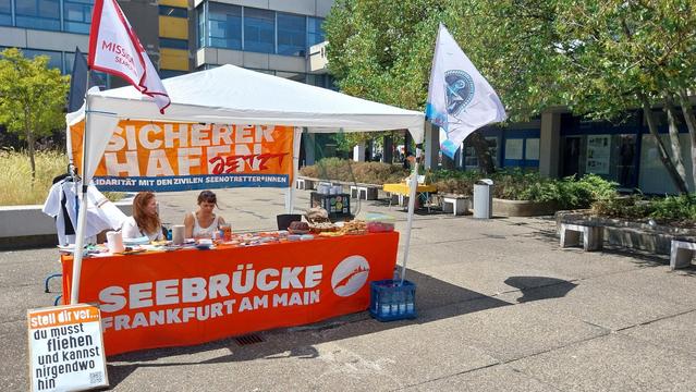 Seebrücke Stand auf dem Campus
 Bockenheim in einem weißen Pavillon.
Über die ganze Breite geht ein Infotisch mit einer orangen Tischdecke, auf der steht in weiß SEEBRÜCKE FRANKFURT AM MAIN und das SB FFM Logo.
An der Rückwand hängt ein oranges Transpi auf dem steht [weiß] SICHERER HAFEN [rot] JETZT, unten in einem blauen Kasten [weiß] SOLIDARITÄT MIT DEN ZIVILEN SEENOTRETTER*INNEN.
Links auf dem Tisch Infomaterial &Sticker, rechts verschiedene Kuchen.
An der rechten Seite des Pavillons ein Tisch mit Dosen, links ein Kleiderständer mit schwarzen & weißen T-Shirts.
Vorne links am Tisch ein Karton mit Plakaten "Stell Dir vor, Du musst fliehen und kannst nirgendwo hin."