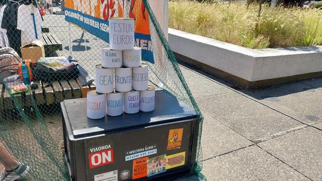 Gestapelte Dosen. Beschriftet mit RASSISMUS, ANTISEMITISMUS, NATIONALISMUS, QUEERFEINDLICHKEIT, FESTUNG EUROPA, PUSHBACKS, GEAS  FRONTEX  ABSCHIEBUNG. BEZAHLKARTE