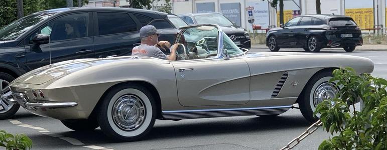 A beige vintage convertible car, possibly a Chevrolet Corvette, is parked on the street. A man with a cap is sitting in the driver's seat, holding the steering wheel. Another car is visible in the background, along with some greenery nearby.