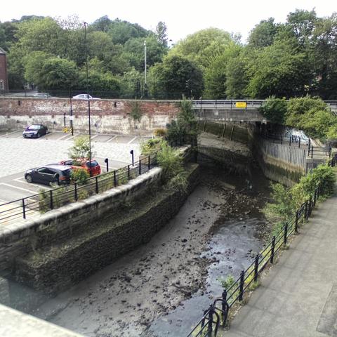 Photograph of the tidal stretch of Ouseburn River at south side of Ouseburn Bridge in Newcastle upon Tyne. To the left (south-west) of the bridge is Cut Bank, and to the right (north-east) is Byker Bank. In the foreground is the pedestrian Riverside Walk and the photo also spans some surrounding built-up areas and trees, with sky in the background. Still image captured from webcam at 17:00 hrs on Saturday 5 July 2025.
