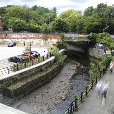 Photograph of the tidal stretch of Ouseburn River at south side of Ouseburn Bridge in Newcastle upon Tyne. To the left (south-west) of the bridge is Cut Bank, and to the right (north-east) is Byker Bank. In the foreground is the pedestrian Riverside Walk and the photo also spans some surrounding built-up areas and trees, with sky in the background. Still image captured from webcam at 18:36 hrs on Saturday 5 July 2025.