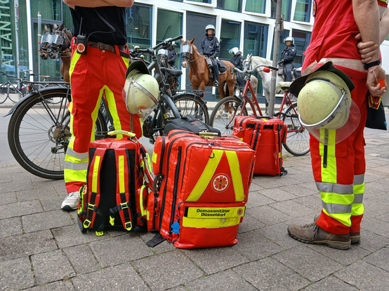 Hier sieht man 2 Demosanitäter*innen von der Hüfte abwärts, sie Tragen Rot,Gelbe Rettungsdienst Hosen und am Gürtel Hängt jewails ein Helm und ein Funkgerät.
Die Beiden Sanis stehen links und rechts vom Bild, in der Mitte stehen 4 Rettungsrucksäcke gesammelt, 3 Große 1 Kleiner. Im Hintergund sieht man Berittene Polizist*innen, die Pferde tragen Schutzvisiere über denn Augen, und die Polizist*innen Volle Schutzausrüstung.