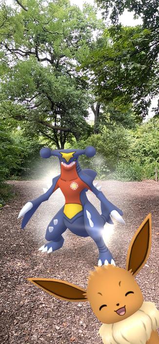 An augmented reality scene featuring two Pokémon characters: a large blue creature with a red and yellow chest, and a small, happy-looking brown creature with large ears. The background depicts a wooded area with trees and a path covered in leaves.