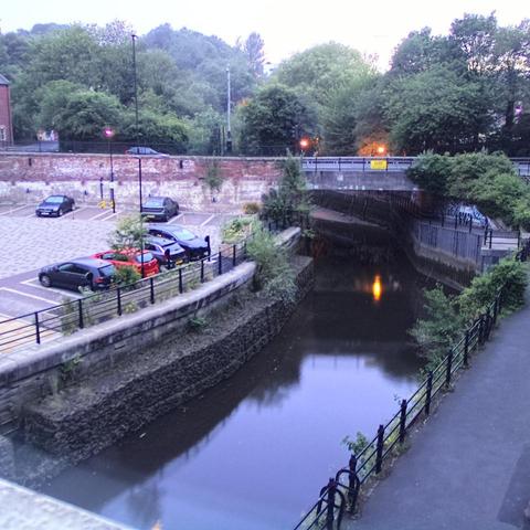 Photograph of the tidal stretch of Ouseburn River at south side of Ouseburn Bridge in Newcastle upon Tyne. To the left (south-west) of the bridge is Cut Bank, and to the right (north-east) is Byker Bank. In the foreground is the pedestrian Riverside Walk and the photo also spans some surrounding built-up areas and trees, with sky in the background. Still image captured from webcam at 21:44 hrs on Saturday 5 July 2025.