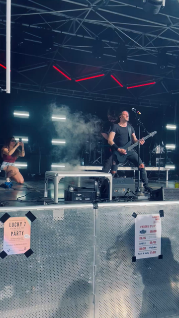 Das Video zeigt eine lebhafte Live-Auftrittszene auf einer Bühne, die von einer Band mit einem intensiven Rock- oder Metal-Stil dominiert wird. Die Bühne ist mit einem modernen Design aus Metallrahmen und roten LED-Lichtern ausgestattet, die eine dynamische Atmosphäre schaffen. Neben dem Hauptgitarrenspieler, der in einem schwarzen Shirt und Rissenhosen steht und eine schwarze Gitarre spielt, ist ein Bassist zu sehen, der eine blaue Bassgitarre spielt. Beide Musiker sind in voller Aktion, mit d…