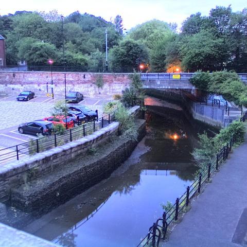 Photograph of the tidal stretch of Ouseburn River at south side of Ouseburn Bridge in Newcastle upon Tyne. To the left (south-west) of the bridge is Cut Bank, and to the right (north-east) is Byker Bank. In the foreground is the pedestrian Riverside Walk and the photo also spans some surrounding built-up areas and trees, with sky in the background. Still image captured from webcam at 04:32 hrs on Sunday 6 July 2025.