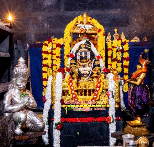 Idol of Hindu deity Krishna in seated in a gold cradle, wearing a necklace with a large lotus-shaped medallion. A small gold umbrella over Krishna's head. Dense string of yellow flowers fall on either side of the umbrella. Strings of white flowers fall from the cradle down to the floor.

A silver figure of Arjuna on his knees, hands folded, in front of Krishna on his left. A bronze idol draped in purple silk cloth, holding a red-orange metallic seal/stamp.

Blue cloth background. Several strings of yellow flowers hang from the platform the cloth covers.