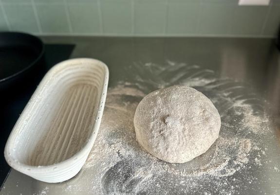 Ein Gärkörbchen steht am linken Bildrand auf der Anrichte aus Chromstahl. Daneben, der rundgewirkte Teigling auf viel Mehl.