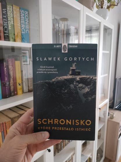 Osoba trzyma książkę pt. Schronisko, które przestało istnieć autorstwa Sławka Gortycha. Na okładce widnieje zdjęcie górskiego schroniska w zimowej scenerii. W tle widoczny regał z książkami oraz wnętrze mieszkania.