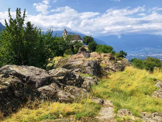 Some rocks and a trail near some trees, a tiny church in the distance and clouds in the sky