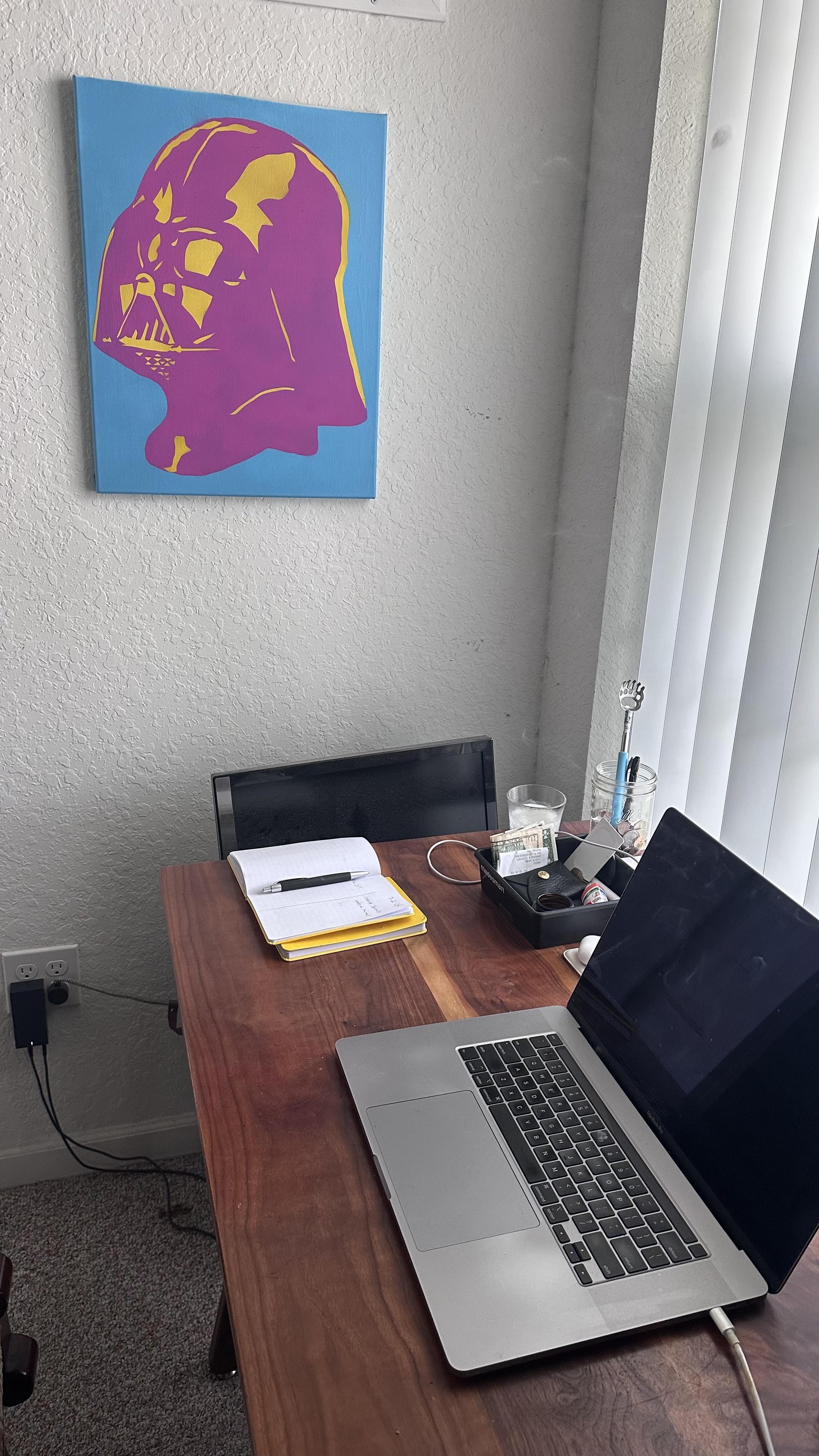 My office space. A colorful blue, white, and pink portrait of a Darth Vader hangs on the wall. It's in the style of pop art. A wooden desk holds a laptop, an open notebook, and a black organizer with various items. Natural light streams in through vertical blinds.