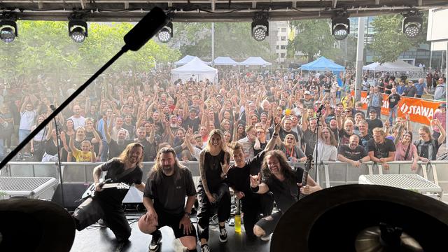 Das Bild zeigt eine Band auf einer Bühne, die vor einer großen, jubelnden Menge steht. Die Bandmitglieder sind in der Mitte des Bildes zu sehen, alle in dunkler Kleidung, einige mit langen Haaren. Sie posieren fröhlich und machen die berühmte "Rock-On"-Geste mit ihren Händen. Die Bühne ist mit Lichtern und Mikrofonen ausgestattet. Im Hintergrund sieht man eine große Menschenmenge, die die Band anfeuert und die Hände hebt. Viele Zuschauer tragen Rock-Kleidung und haben Gesichter, die Freude und …