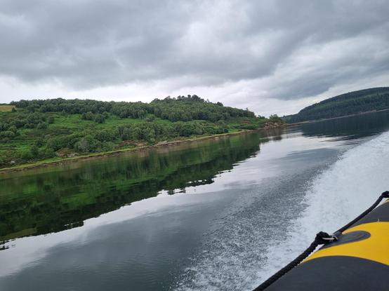 Looking back from aboard a speedboat you see a tiny bit of the boat and some whitewater behind it. The rest is just water, green, Scottish hills, and a very cloudy, Scottish sky.