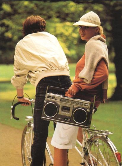 Couple on a bicycle, carrying a boombox, in a park setting.