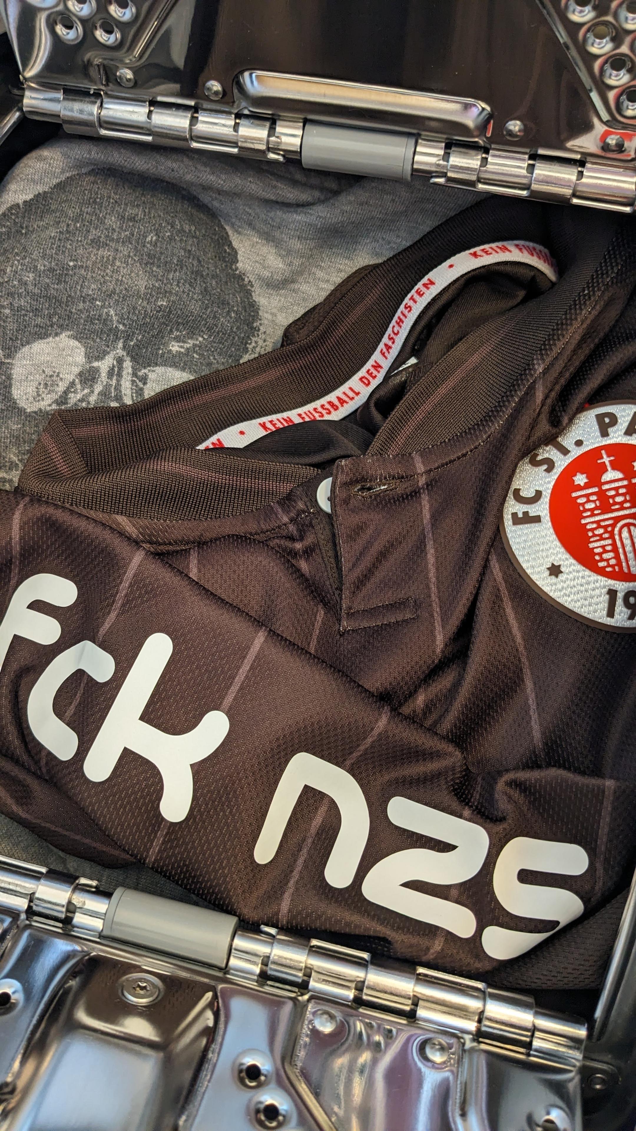 Bildbeschreibung von KI:
Das Foto zeigt den geöffneten Koffer aus silberfarbenem Metall mit glänzender Oberfläche und sichtbaren Scharnieren. Im Koffer liegt ein zusammengefaltetes braunes Trikot des Fußballvereins FC St. Pauli. Auf dem Trikot ist das Vereinswappen gut erkennbar. Es zeigt in einem roten Kreis ein stilisiertes weißes Stadttor mit der Jahreszahl neunzehnhundert zehn. Daneben ist in großen weißen Buchstaben die Abkürzung fck nzs aufgedruckt, eine politische Botschaft gegen Nationalsozialismus. Am inneren Kragenrand des Trikots steht in roter Schrift der Satz kein Fußball den Faschisten. Unter dem Trikot ist ein graues T-Shirt mit einem Totenkopf-Motiv zu sehen, das Symbol des Vereins und seiner Fanszene. Die Szene wirkt ordentlich und bewusst zusammengestellt, mit klarer politischer Haltung und Fußballbezug.
Bildbeschreibung von KI:
Das Foto zeigt den geöffneten Koffer aus silberfarbenem Metall mit glänzender Oberfläche und sichtbaren Scharnieren. Im Koffer liegt ein zusammengefaltetes braunes Trikot des Fußballvereins FC St. Pauli. Auf dem Trikot ist das Vereinswappen gut erkennbar. Es zeigt in einem roten Kreis ein stilisiertes weißes Stadttor mit der Jahreszahl neunzehnhundert zehn. Daneben ist in großen weißen Buchstaben die Abkürzung fck nzs aufgedruckt, eine politische Botschaft gegen Nationalsozialismus. Am inneren Kragenrand des Trikots steht in roter Schrift der Satz kein Fußball den Faschisten. Unter dem Trikot ist ein graues T-Shirt mit einem Totenkopf-Motiv zu sehen, das Symbol des Vereins und seiner Fanszene. Die Szene wirkt ordentlich und bewusst zusammengestellt, mit klarer politischer Haltung und Fußballbezug.