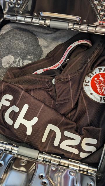 Bildbeschreibung von KI:
Das Foto zeigt den geöffneten Koffer aus silberfarbenem Metall mit glänzender Oberfläche und sichtbaren Scharnieren. Im Koffer liegt ein zusammengefaltetes braunes Trikot des Fußballvereins FC St. Pauli. Auf dem Trikot ist das Vereinswappen gut erkennbar. Es zeigt in einem roten Kreis ein stilisiertes weißes Stadttor mit der Jahreszahl neunzehnhundert zehn. Daneben ist in großen weißen Buchstaben die Abkürzung fck nzs aufgedruckt, eine politische Botschaft gegen Nationalsozialismus. Am inneren Kragenrand des Trikots steht in roter Schrift der Satz kein Fußball den Faschisten. Unter dem Trikot ist ein graues T-Shirt mit einem Totenkopf-Motiv zu sehen, das Symbol des Vereins und seiner Fanszene. Die Szene wirkt ordentlich und bewusst zusammengestellt, mit klarer politischer Haltung und Fußballbezug.
