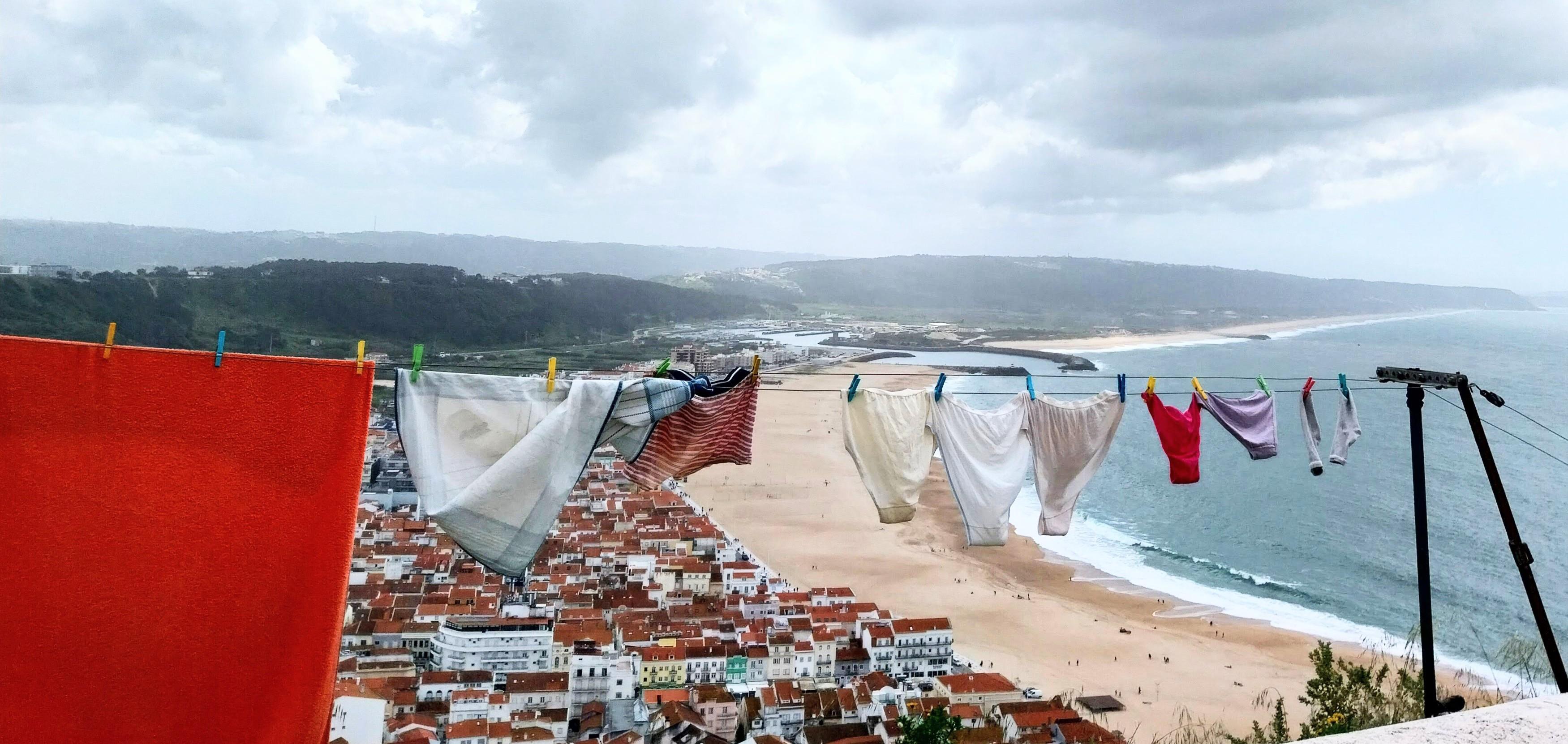 Im Vordergrund sieht man Wäsche, die im Wind weht auf einer Leine. Darunter, sehr klein, die Häuser einer Stadt und davor ein breiter weißer Strand und das Meer. Weit entfernt bewaldete Hügel. Der Himmel stark bewölkt Im Vordergrund sieht man Wäsche, die im Wind weht auf einer Leine. Darunter, sehr klein, die Häuser einer Stadt und davor ein breiter weißer Strand und das Meer. Weit entfernt bewaldete Hügel. Der Himmel stark bewölkt