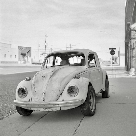 A black and white film photo. An old Volkswagen Beetle, who has a missing windshield, headlights and is dented up all over, sits quietly on a sidewalk in Lubbock, Texas. Sunday, February 16, 2025.