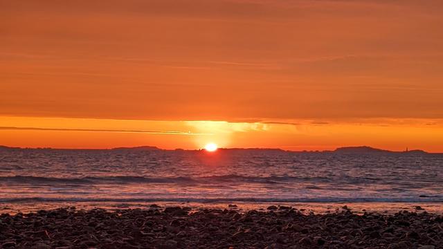 Historical Documentation in the form of a photograph of a rising sun over the ocean bay at Yirrell Beach in Winthrop, MA. The sky is filled with vibrant orange and yellow hues, with the sun partially visible in the center of the image, just behind a distant island. The sun is rising over the horizon, casting a warm glow over the water and the horizon as it sets the sky a blaze in a gradient of orange hues. The water is calm as the waves gently crash onto the shore. In the foreground, there is a rocky beach with small pebbles scattered across it. On the right side of the image, there are a few small islands visible. The overall mood of the photograph is peaceful and serene as we witness the dawn of a new day.