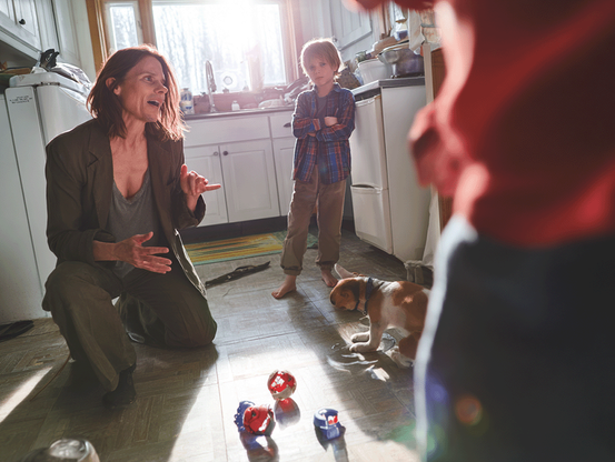 A woman kneels on the kitchen floor, gesturing passionately as she speaks to a child, suggesting a serious or emotional conversation. A young boy stands barefoot in the background with his arms crossed and a tense expression, while a small dog nearby sniffs the floor. Toys are scattered across the floor, and another child in a red shirt is partially visible in the foreground, slightly out of focus. Sunlight streams through the window, casting a warm glow across the scene. The kitchen appears cluttered and lived-in.