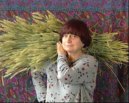 Filmstil aus dem Film LES GLANEURS ET LA GLANEUSE von Agnès Varda. Zu sehen ist Agnès Varda vor einem bunten Tuch mit frisch geschnittenem Getreide auf der rechten Schulter.