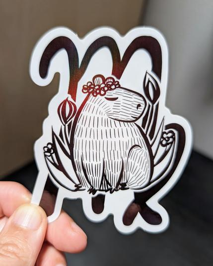 Photo of a left hand holding a diecut sticker. The sticker is a holographic drawing of a capybara Virgo zodiac sign.