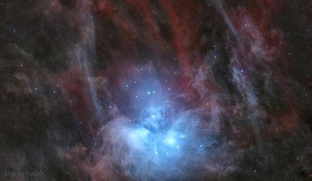 A wide-field astrophotograph of the Pleiades star cluster (M45) shows brilliant blue stars surrounded by glowing blue nebulosity. Wisps of dust appear to stretch like mist around the cluster. A soft red glow from hydrogen gas surrounds the outer edges, creating a vivid contrast against the dark backdrop of space.