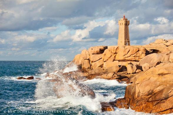 Leuchtturm steht auf Fels, im Vordergrund schlägt eine Welle gegen einen Stein