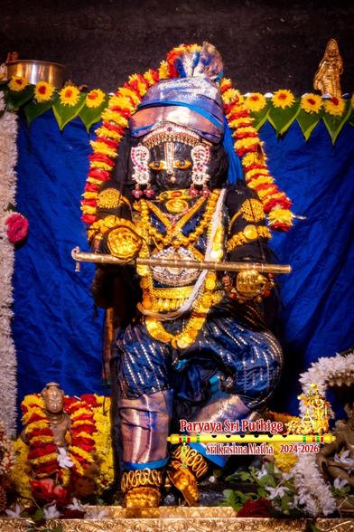Idol of Hindu deity Krishna standing dressed in bright royal blue and a matching crested turban. Left leg bent slightly at the knee for a casual appearance ("contrapposto"). Wearing gold shoes, gold palm coverings, and several gold ornaments including a large necklace made of gold coins. Holding a gold flute in hands and standing on a gold-covered slab. A string of alternating yellow and red flowers shoulder-shoulder going over the turban. Blue cloth background with a string of green beetle leaves at the top.
