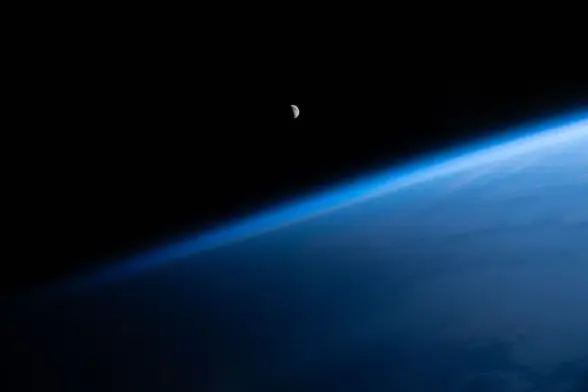 A photograph of the distant waning crescent moon, lit from the right, seen over the Atlantic Ocean. The blackness of space serves as the background, and the blue haze of the atmosphere is visible between the Earth and space.