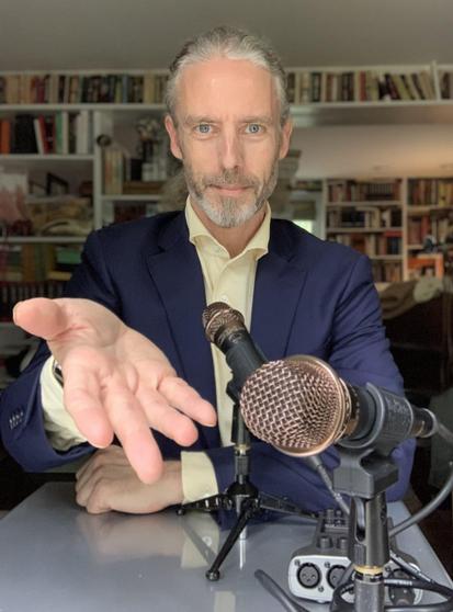 Photo of bearded white man in blue suit & light yellow longsleeved poll sitting at a small silver table with a pair of microphones on tripods feeding into a digital audio recorded. He's gesturing to the viewer to sit down and gab.
