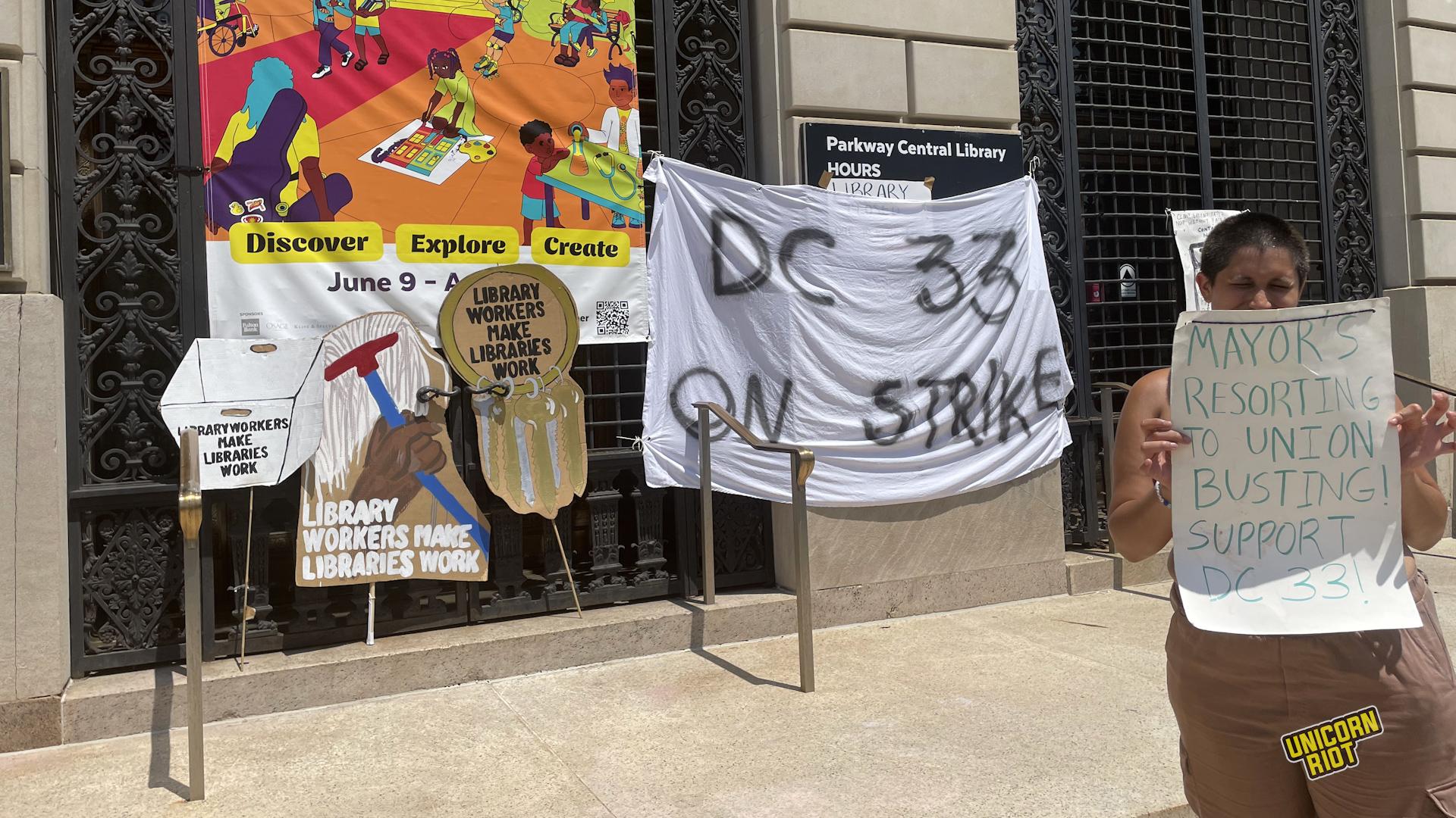 Union signs banners and signs lean against the front of the Parkway library branch as a striker or supporter holds a hand-written sign reading "MAYOR'S RESORTING TO UNION BUSTING! SUPPORT DC 33!"