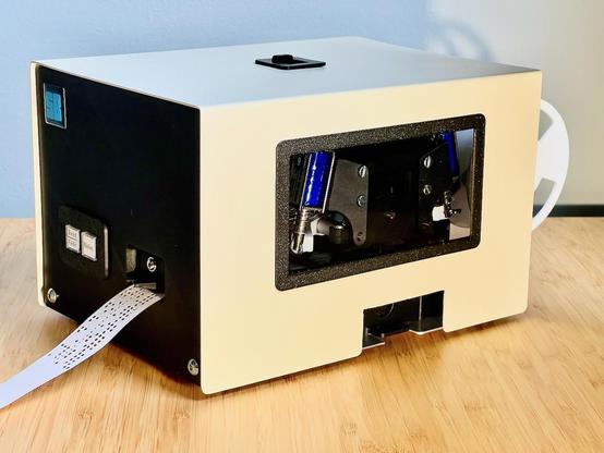 A beige box with a black end and a paper spool on the other side,  a window into the inside and a paper tape with structured holes coming out the other end
